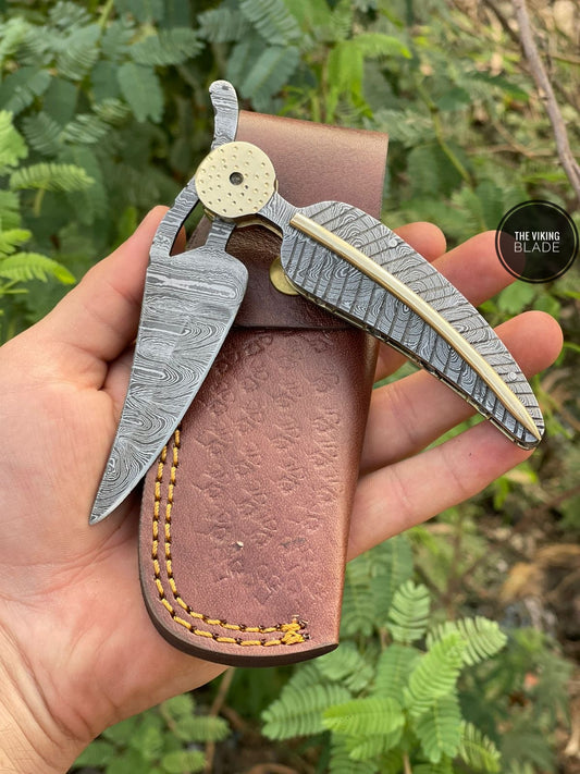 A hand holding a folded Damascus folding knife with Damascus brass file work, alongside a brown leather sheath with decorative stitching.