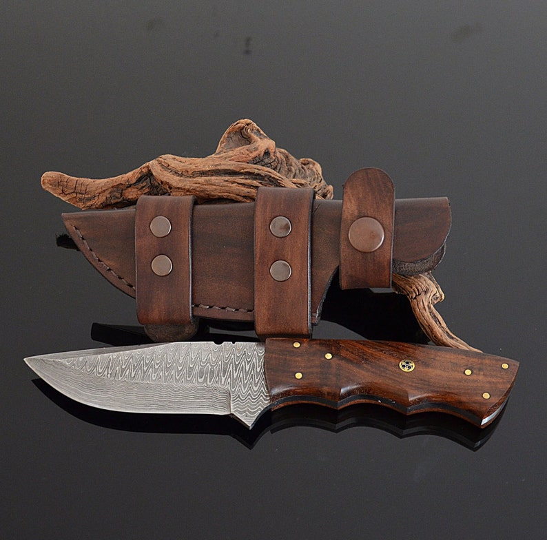 Hand-forged Damascus hunting knife with a brown wooden handle and brass accents, accompanied by a brown leather knife sheath.