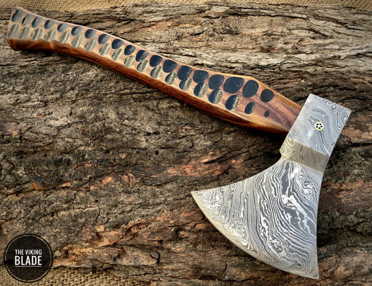 A Damascus steel axe with a patterned blade and a smooth, dark wooden handle, resting on a natural wood surface.