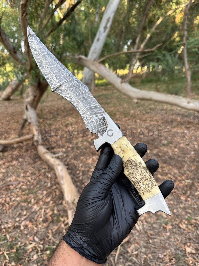 Hand holding a knife with a detailed blade and wooden handle against a forest background