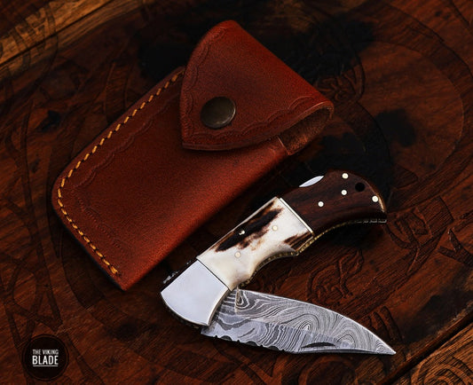 A folding pocket knife with a Damascus steel blade and a brown leather sheath. The knife is open, displaying the blade and the handle, which has a dark, textured finish.
