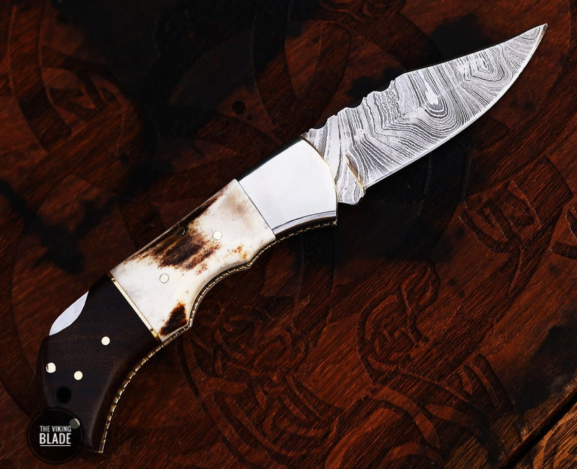 A folding pocket knife with a Damascus steel blade and a brown leather sheath. The knife is open, displaying the blade and the handle, which has a dark, textured finish.