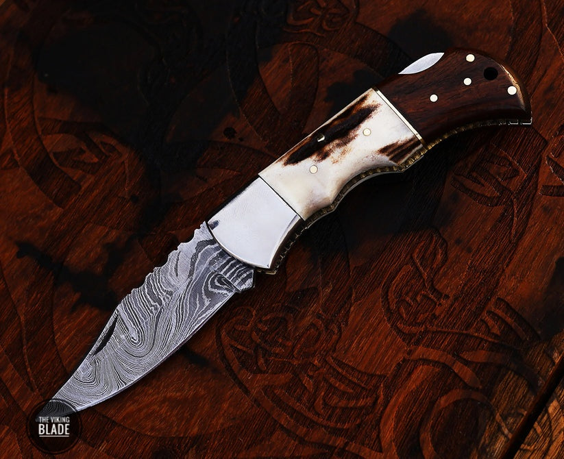 A folding pocket knife with a Damascus steel blade and a brown leather sheath. The knife is open, displaying the blade and the handle, which has a dark, textured finish.