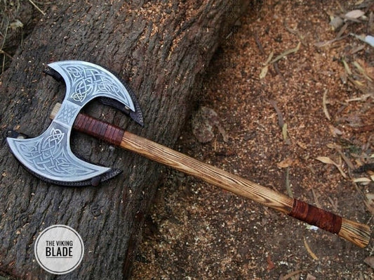 A Viking axe with a double blade featuring Norse patterns, a wooden handle, and a leather sheath, placed against a natural outdoor background.