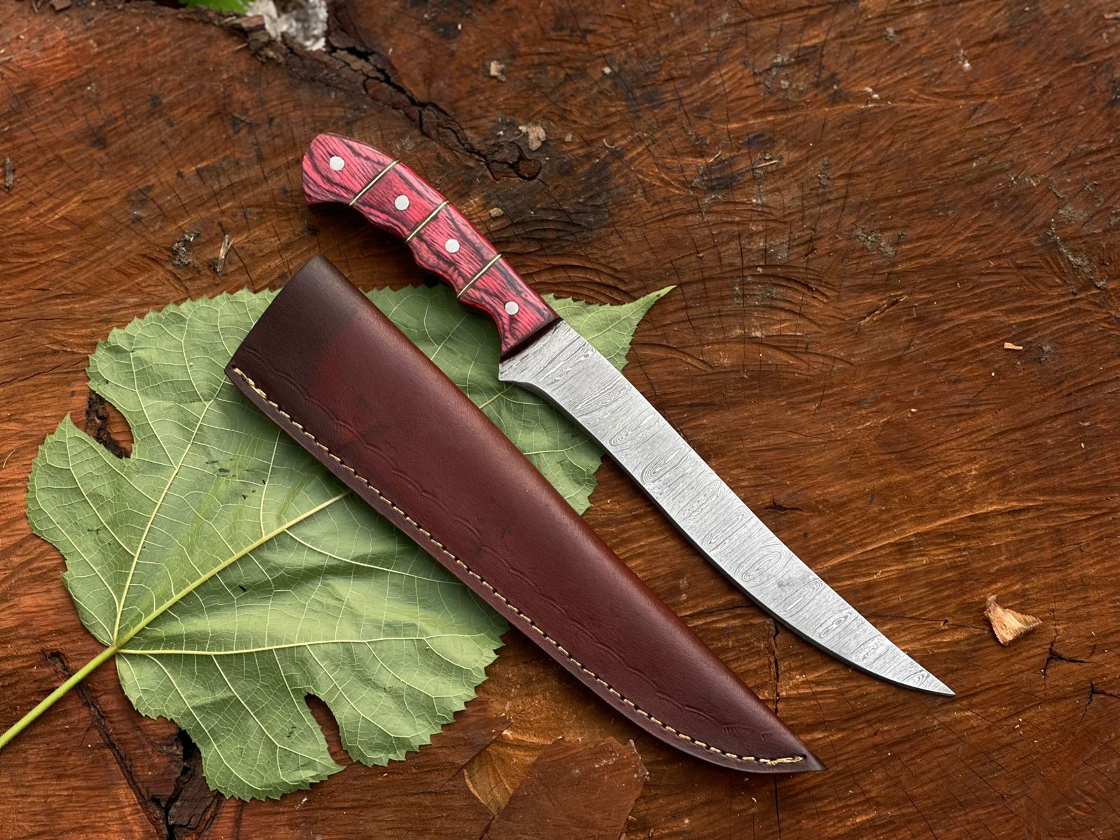 Knife with red handle and brown sheath on a wooden surface with a leaf