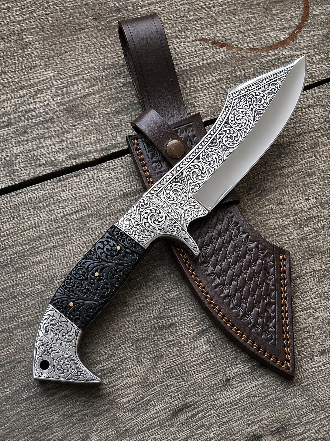 An ornate fixed-blade knife with a silver engraved blade, black and silver handle, and a brown leather sheath, displayed diagonally on a weathered gray wood surface.