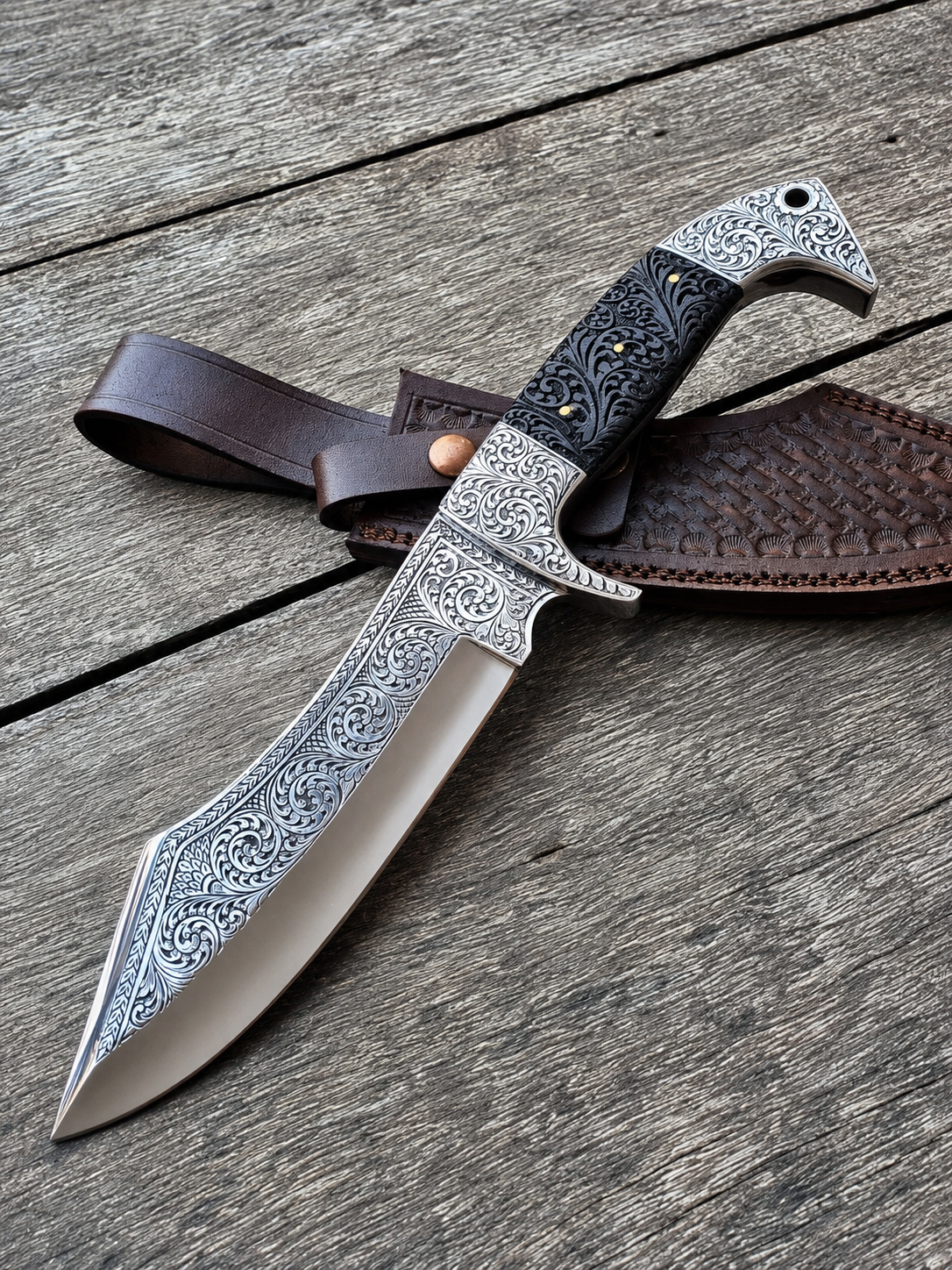 An ornate fixed-blade knife with engraved silver-toned metal detailing, a black handle, and a brown leather sheath, arranged diagonally on a weathered gray wooden surface.