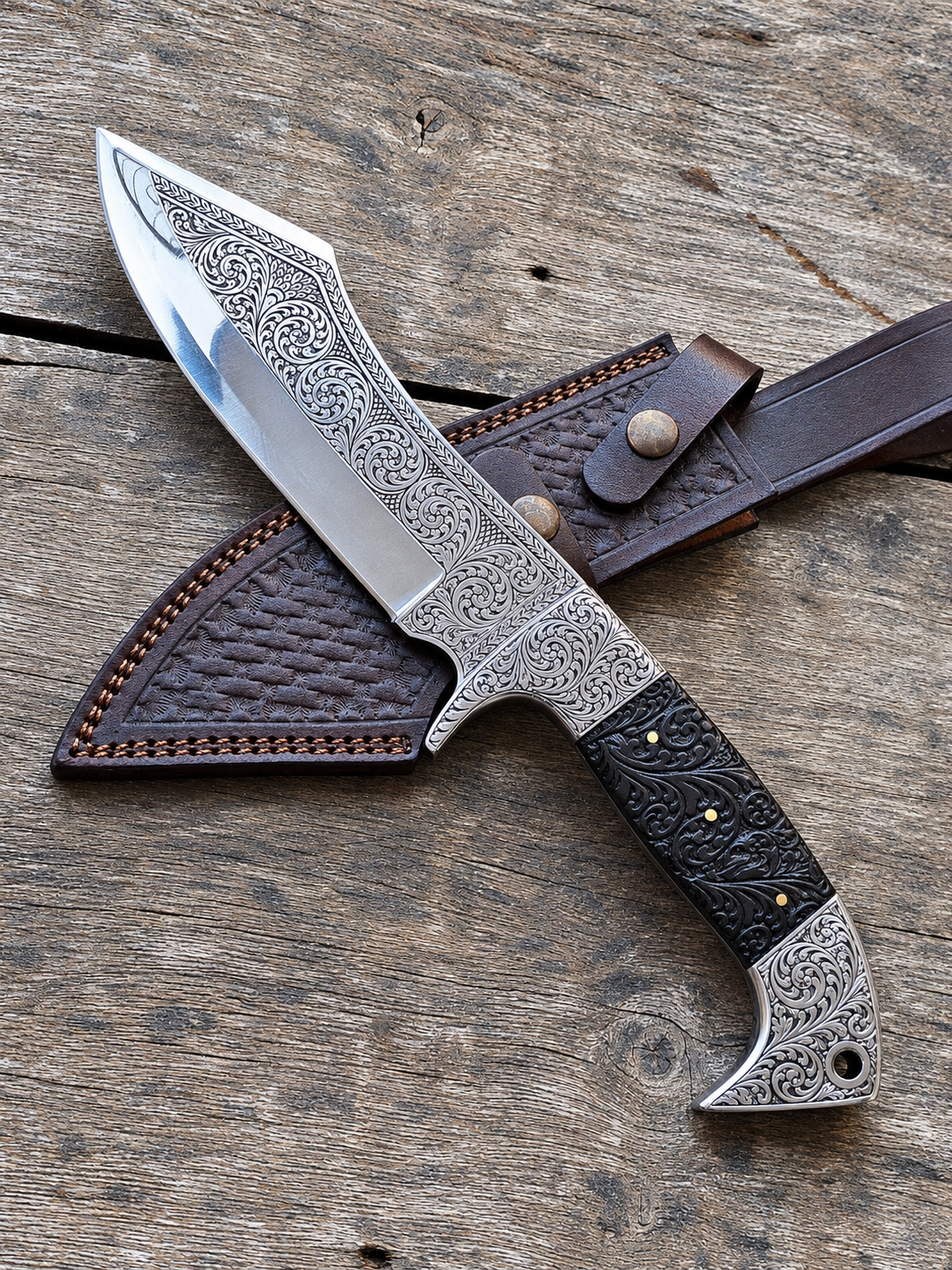 An ornate fixed-blade knife with an engraved silver blade and black handle, resting on a dark brown leather sheath over a rustic wooden surface.