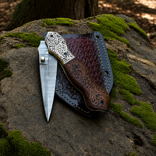 A folding pocket knife with a Damascus steel blade and ornate silver handle rests beside its dark brown leather sheath on a mossy tree root in a forest setting.
