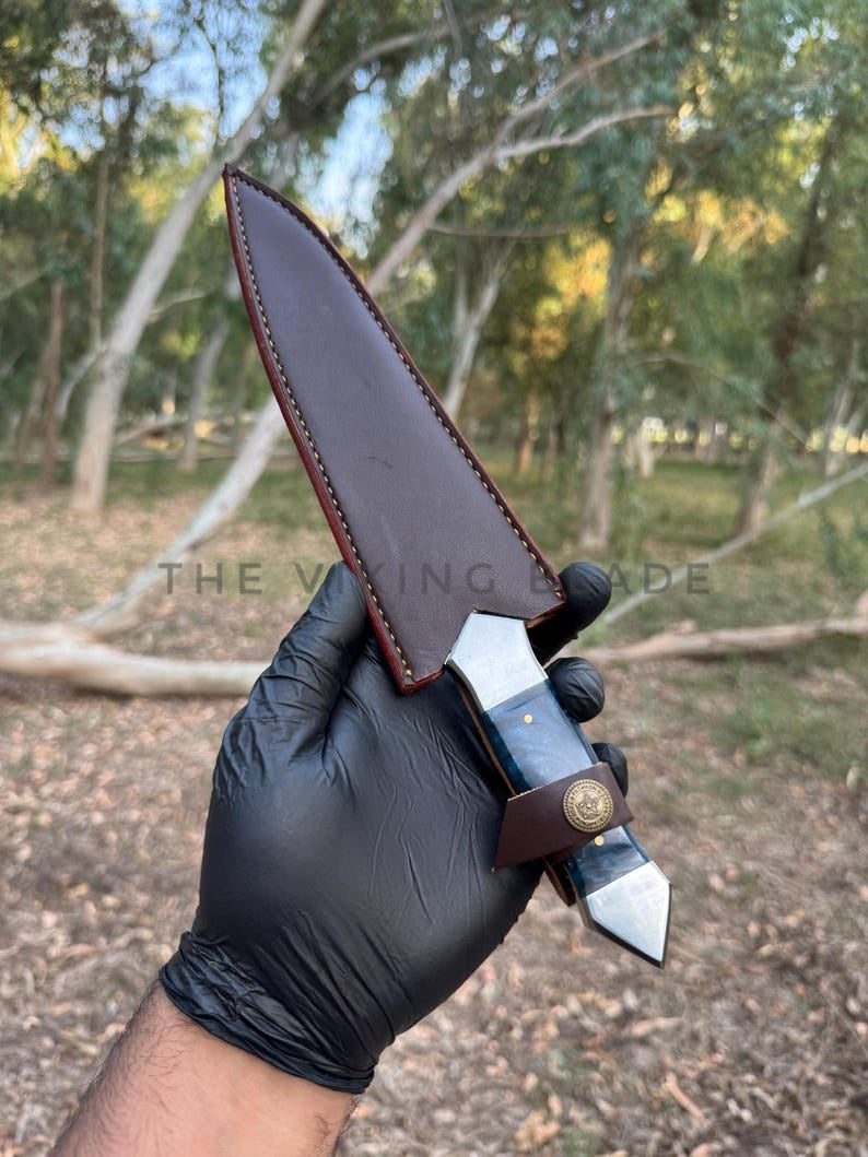 Knife with leather sheath held by a gloved hand in an outdoor setting
