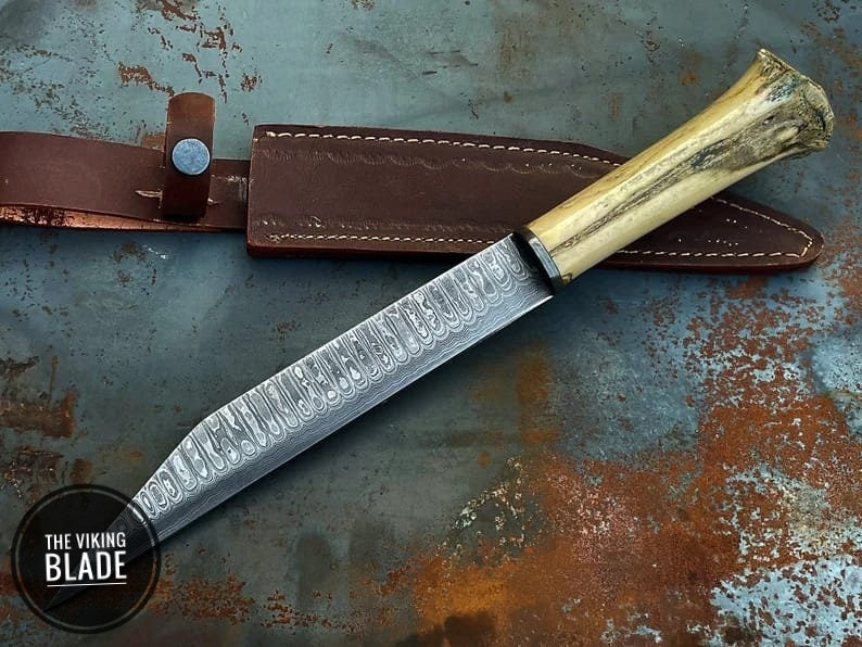 A hand forged stainless steel seax knife with a Damascus pattern blade and a camel bone grip, displayed next to its brown leather sheath, placed on a rusty metal surface.