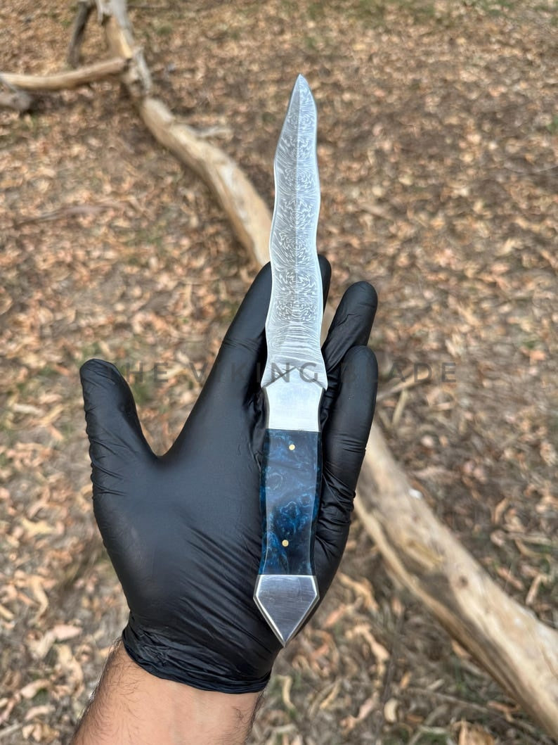 Knife with a blue handle held by a hand wearing a black glove, against a natural background.