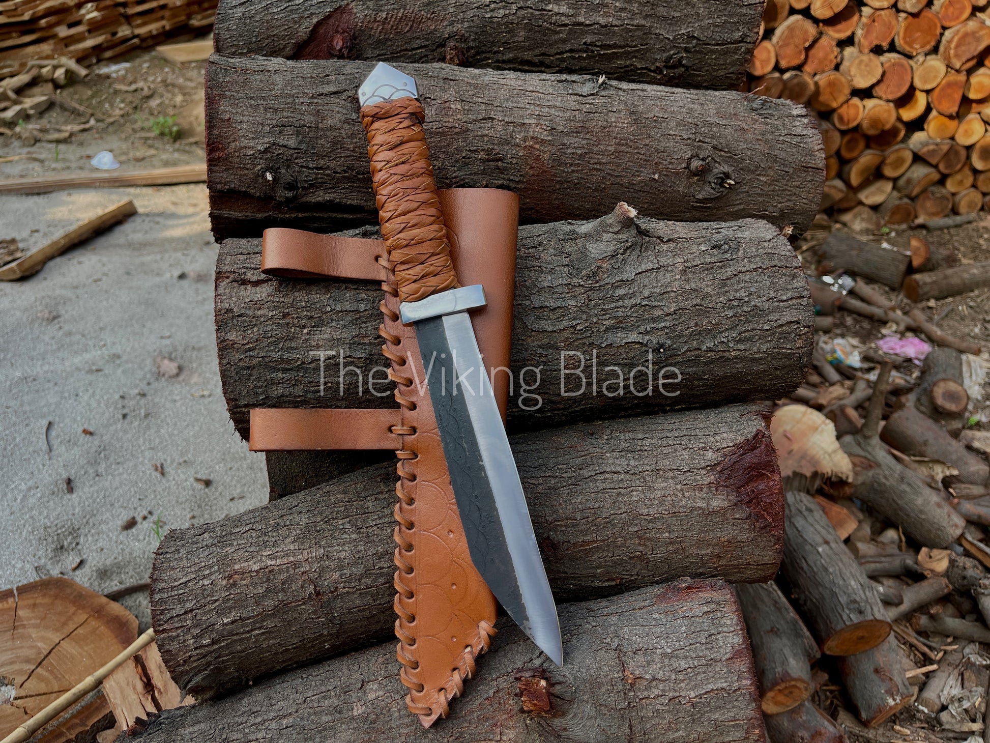 A hand forged troll seax knife with a brown leather sheath, resting on top of a pile of wooden logs.