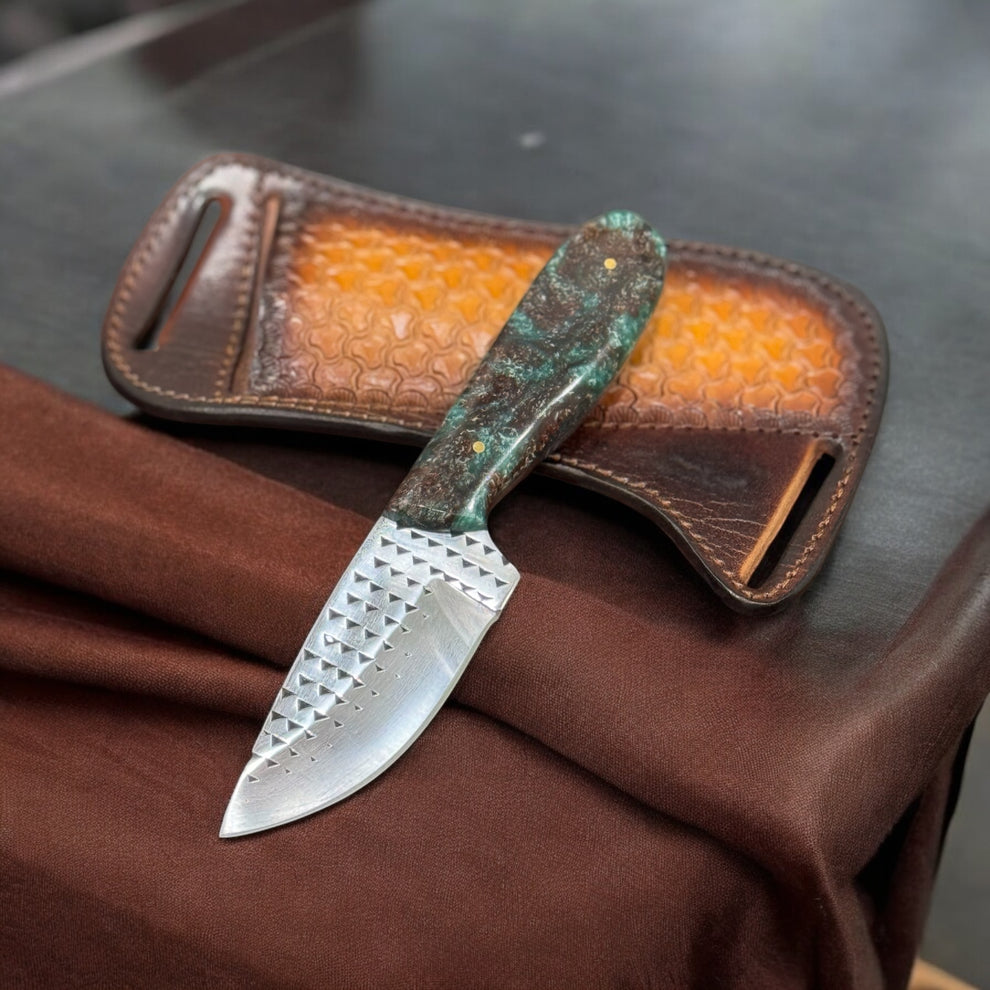 A handcrafted cowboy belt knife with a green and brown handle, placed on a brown surface, with a genuine leather loop case in the background.