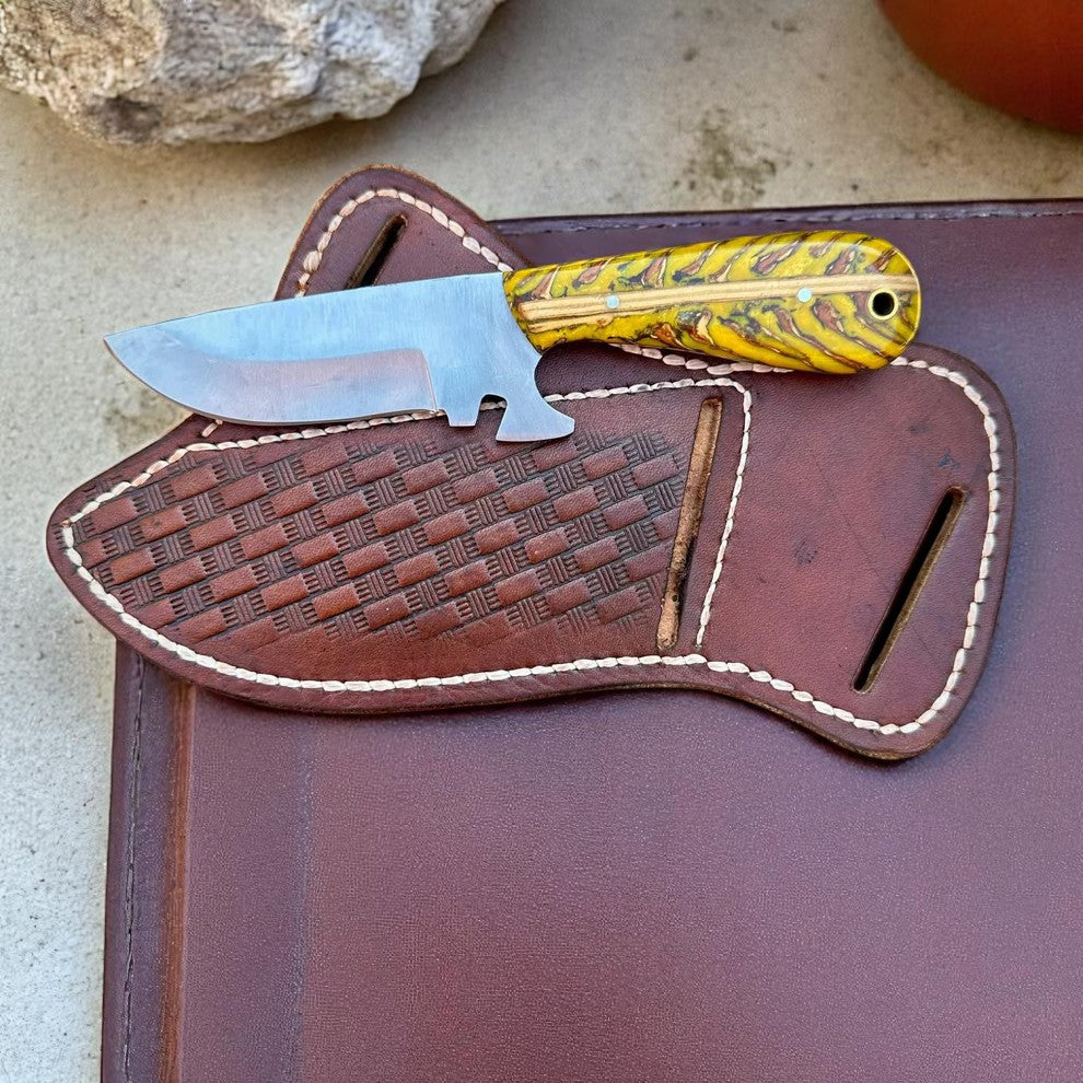 A knife with a J2 steel blade and a resin pine cone handle, placed on top of a brown leather belt sheath. The knife has a partially serrated edge and a brown handle with a natural pine cone design.