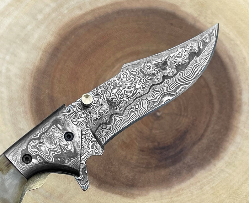 A Damascus steel folding pocket knife with a ram horn handle, displayed alongside its leather pouch.