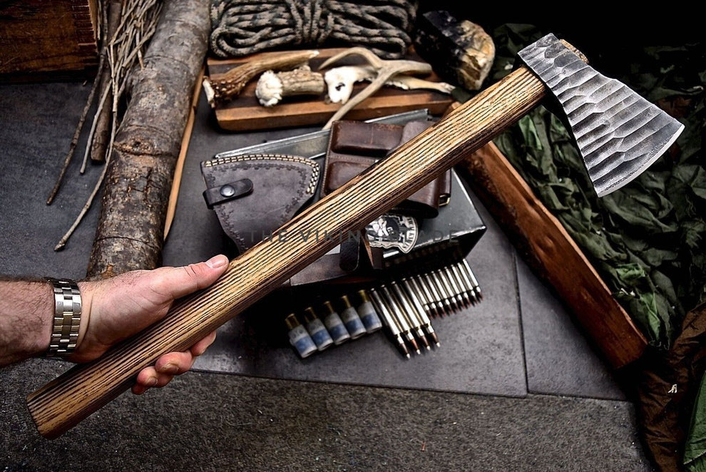 Hand holding a wooden-handled axe over a rustic setting with tools and leather goods.