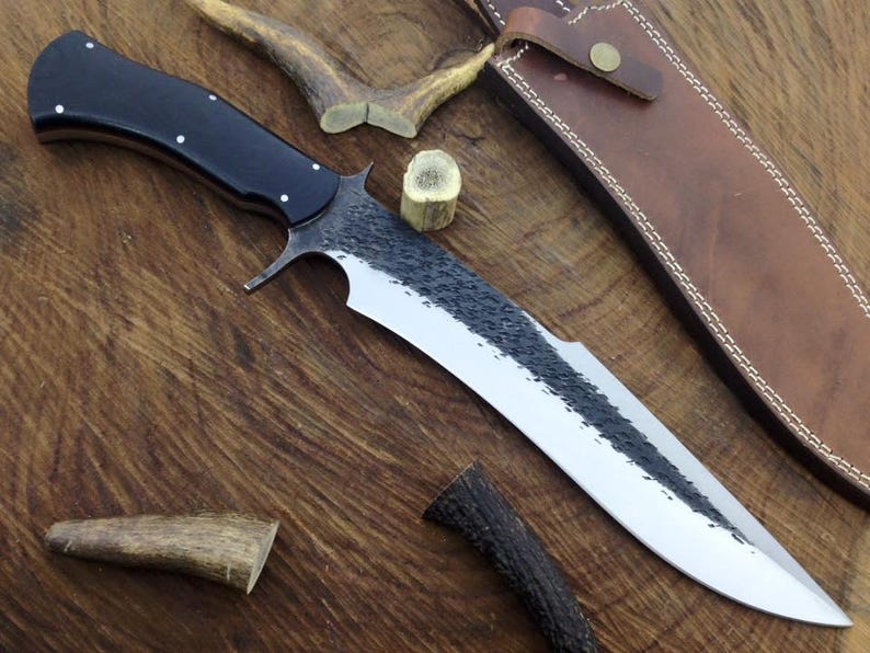 Long knife with a black handle on a wooden surface, accompanied by a leather sheath and antlers.