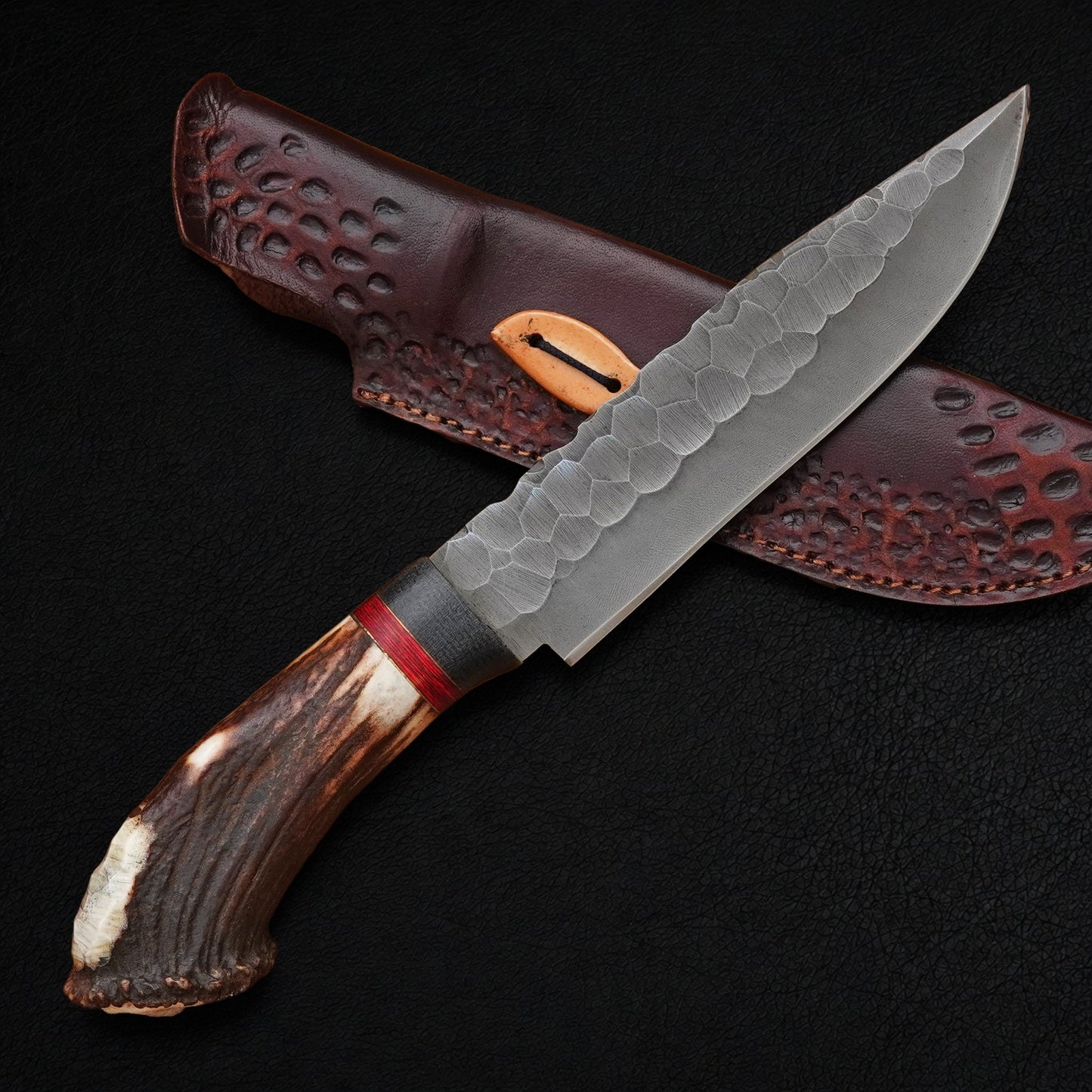 A fixed-blade knife with a hammered steel blade, a brown antler-style handle, and a red accent band, shown diagonally across a dark brown textured leather sheath on a black background.
