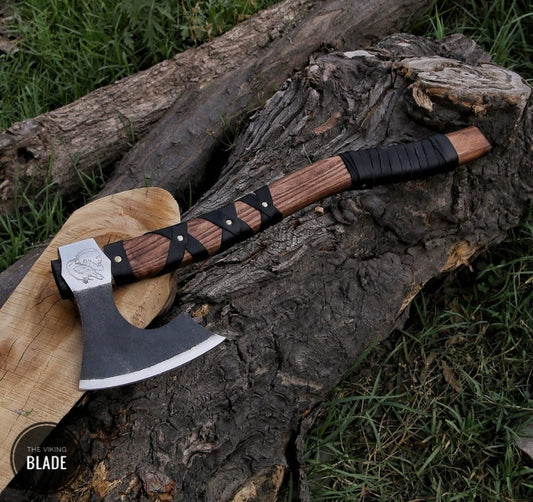 A handmade carbon steel Viking axe with a brown leather-wrapped handle and a black steel blade, resting on a wooden stump with a natural outdoor background.