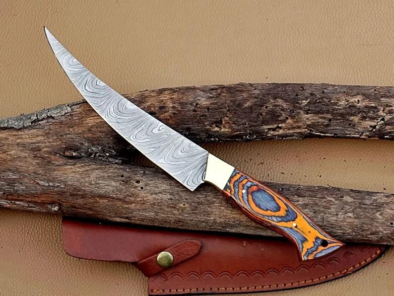 A Damascus steel fillet fishing knife with a brass bolster and pakka wood handle, displayed on a wooden surface with a leather sheath beside it.