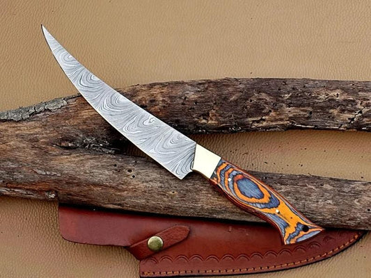 A Damascus steel fillet fishing knife with a brass bolster and pakka wood handle, displayed on a wooden surface with a leather sheath beside it.