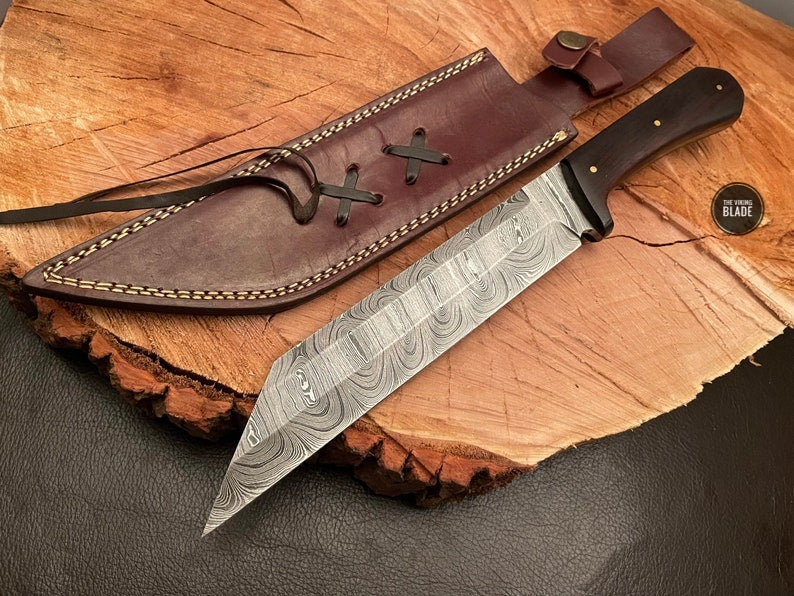A handmade Damascus steel seax with a patterned blade and a brown leather sheath, displayed on a wooden cross-section.