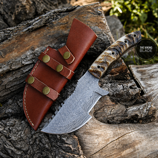 A fixed-blade knife with a patterned steel blade and a carved handle, shown resting on a log next to a brown leather sheath. A circular logo in the upper right reads 'THE VIKING BLADE'.