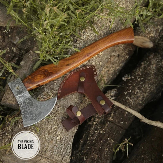 A handmade Damascus Viking axe with a brown wooden handle and a leather sheath, placed on a natural wood surface.