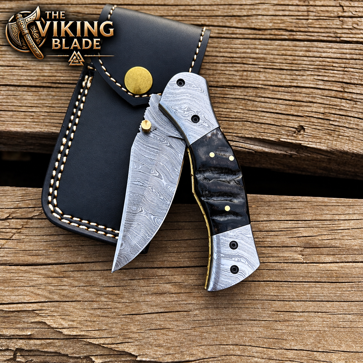 A folding pocket knife with a patterned steel blade and black handle lies on a rustic wooden surface next to a black leather sheath. The logo 'THE VIKING BLADE' is visible in the top-left corner.