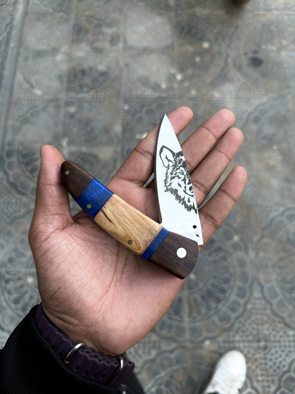 Hand holding a folding knife with a wooden handle and blue accent on a textured surface.
