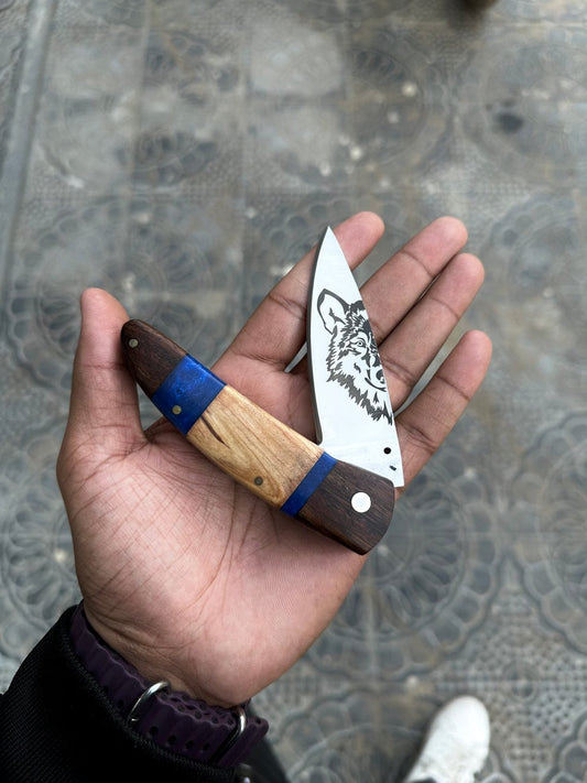 Hand holding a folding knife with a wooden handle and blue accent on a textured surface.
