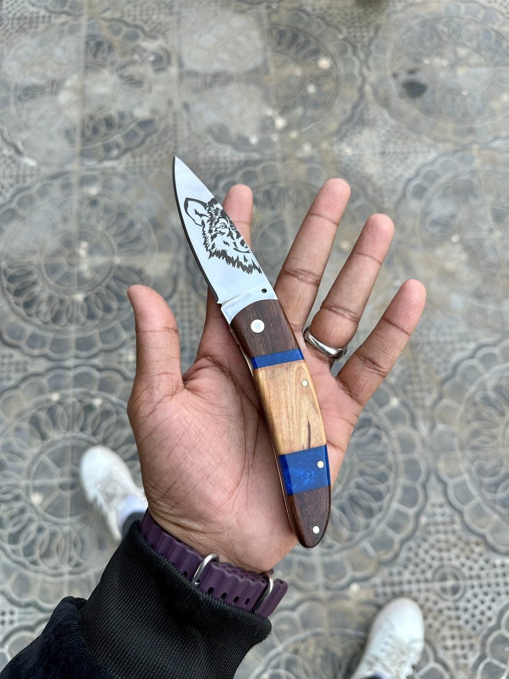 Hand holding a knife with a decorative blade and wooden handle on a textured pavement background