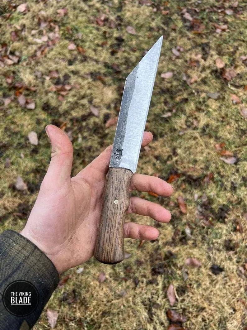 Hand forged Viking knife with a steel blade and wooden handle, held in a hand with a leather sheath visible in the background.