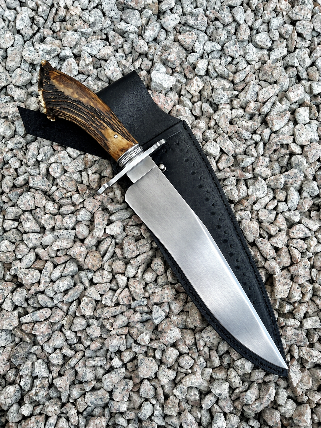 A fixed-blade knife with a brushed silver steel blade, a brown antler-style handle, and a black leather sheath, laid diagonally on a bed of gray gravel.