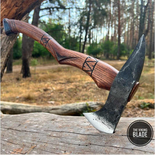 A handmade tomahawk axe with a black head and a brown ashwood handle, displayed outdoors with a leather cover next to it.