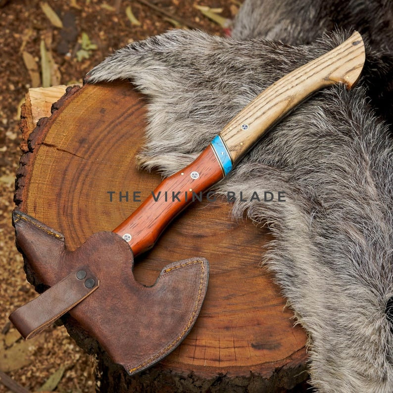Axes with wooden handles and leather sheaths on a wooden log and fur background, featuring 'The Viking Blade' branding.