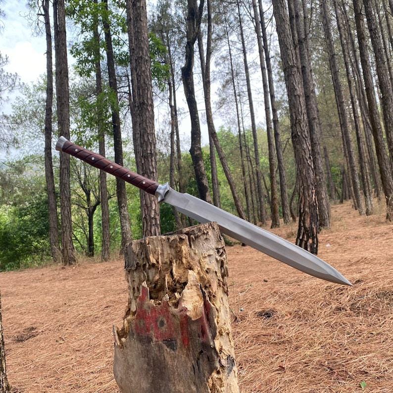 A large fixed-blade knife with a silver blade and a dark brown handle rests on a tree stump in a forest setting with tall pine trees and brown pine needles on the ground.