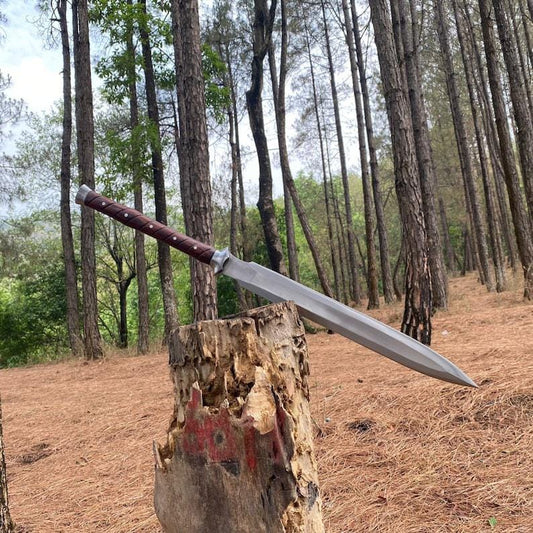 A large fixed-blade knife with a silver blade and a dark brown handle rests on a tree stump in a forest setting with tall pine trees and brown pine needles on the ground.