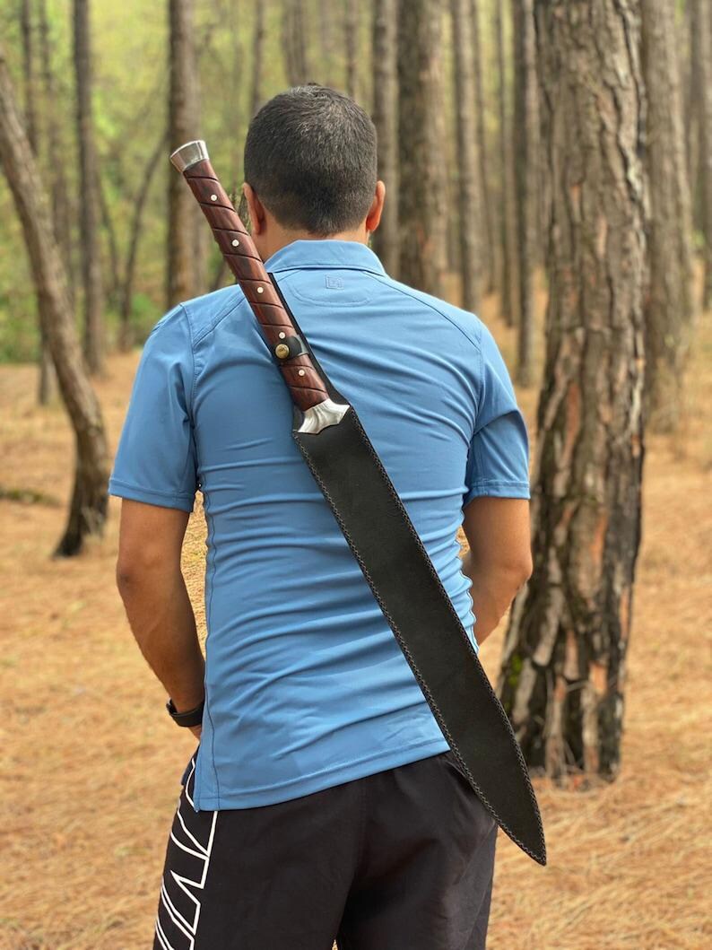 A man viewed from behind in a pine forest, wearing a blue polo shirt and black shorts, with a large machete sheathed on his back.