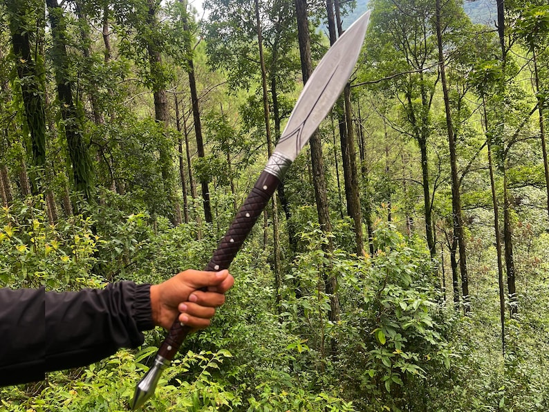 A person's hand holding a long, curved sword with a silver blade, dark brown wrapped handle, and silver metal fittings, set against a dense green forest background.