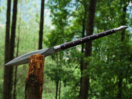 A Viking-style bearded axe with a silver blade and a dark brown wrapped handle, shown embedded in a wooden log with a blurred forest background.