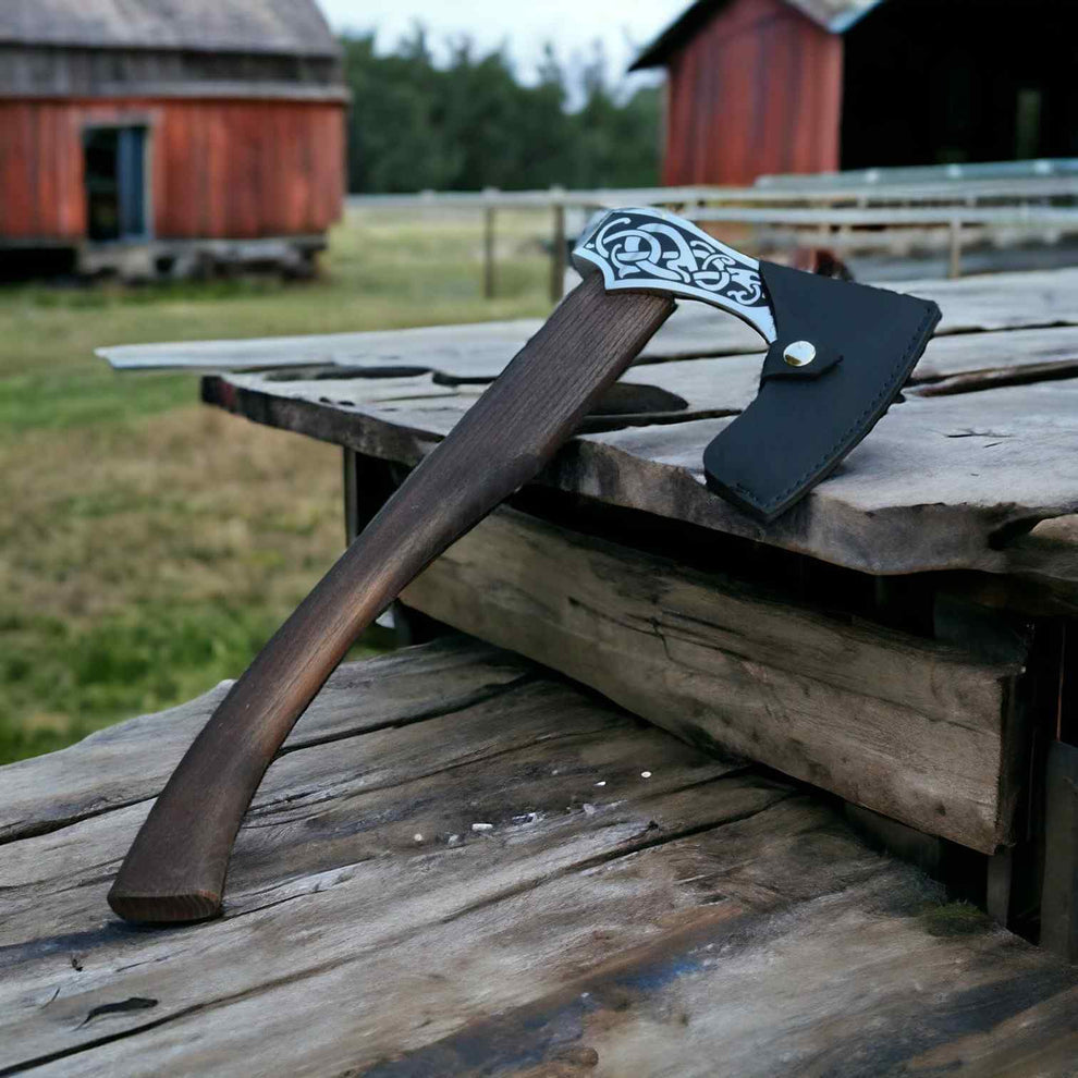 A handcrafted Viking-style axe with a dark wooden handle, ornate silver blade, and black leather sheath, resting on a rustic wooden table with a farm background.