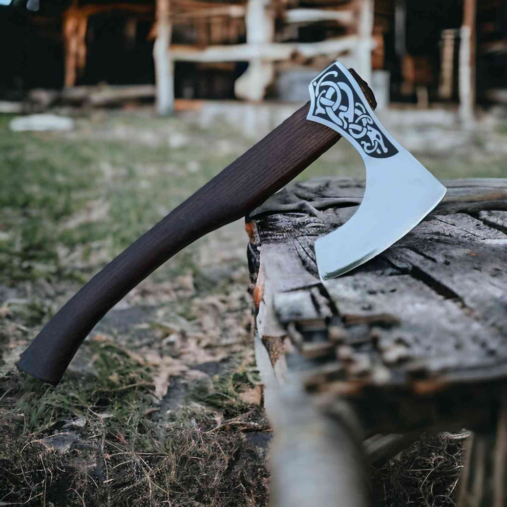 A hand axe with a dark brown wooden handle and a silver steel head featuring a black Norse knotwork pattern, resting on a weathered log in an outdoor rustic setting.