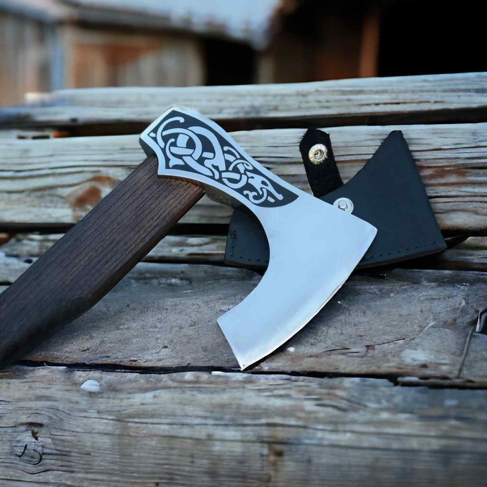 A hand-crafted Viking-style axe with a dark wood handle, ornate silver guard, and polished steel head, resting on weathered wooden planks next to its black leather sheath.