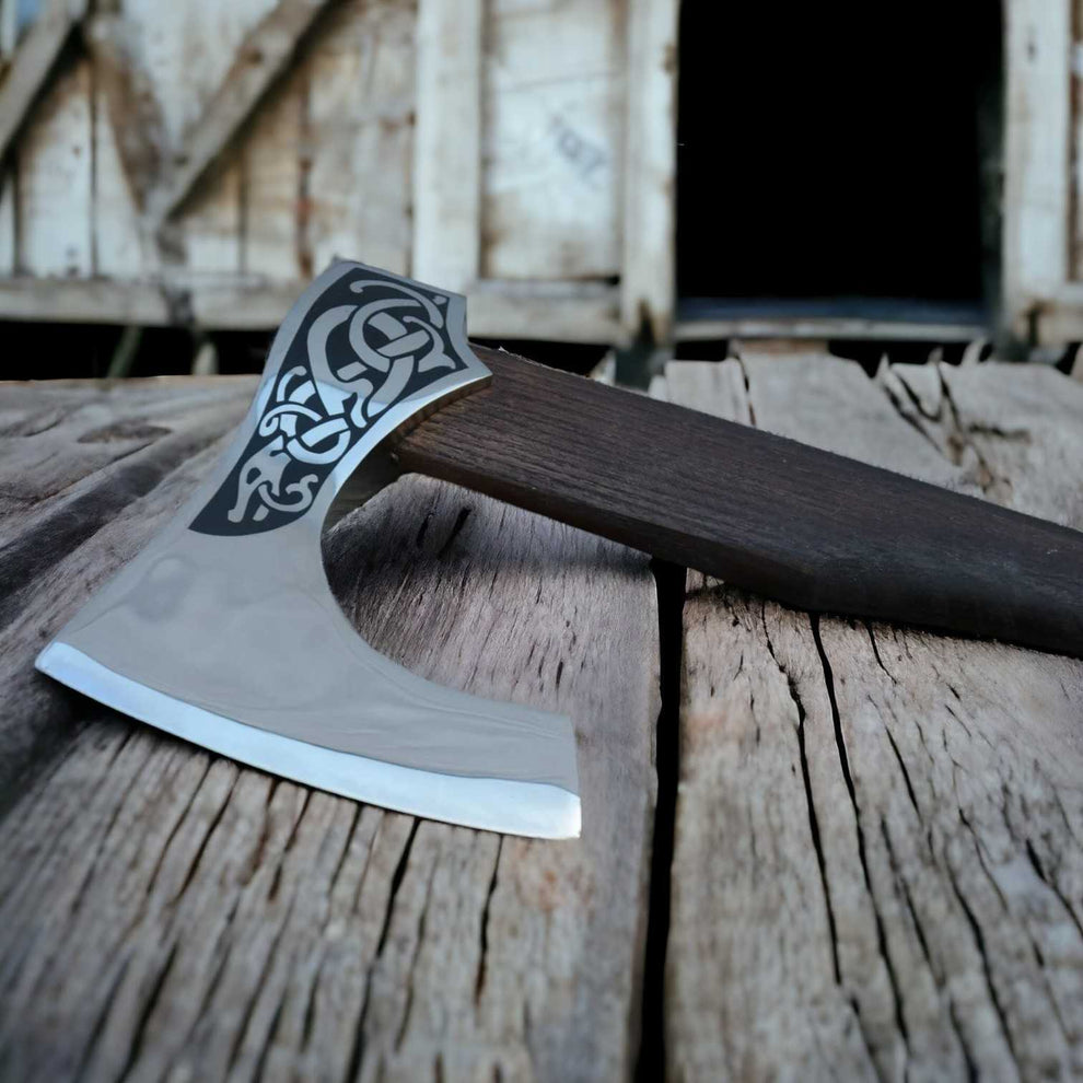 A close-up of a Viking-style bearded axe resting on weathered wooden planks, with a rustic wooden structure blurred in the background.