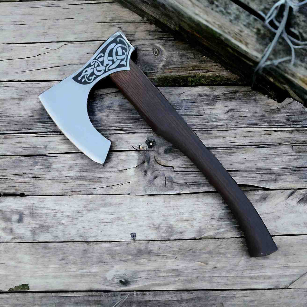 A hand-crafted Viking-style axe with a polished steel blade, ornate black Celtic knot engraving, and a dark brown wooden handle, resting on weathered gray wooden planks.