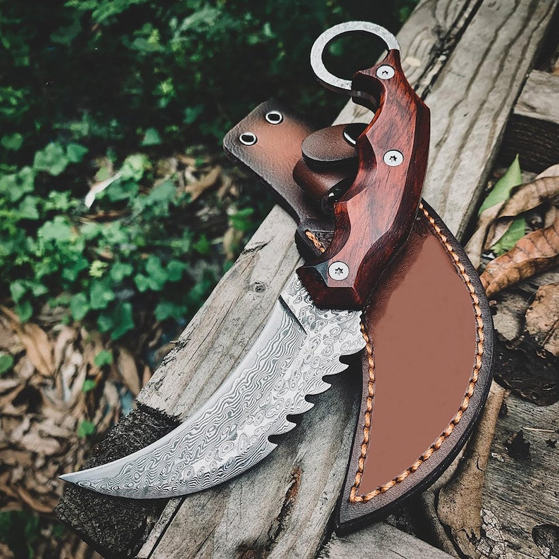 A fixed-blade knife with a patterned steel blade, reddish-brown wooden handle, and brown leather sheath, resting on weathered wood outdoors with green foliage in the background.