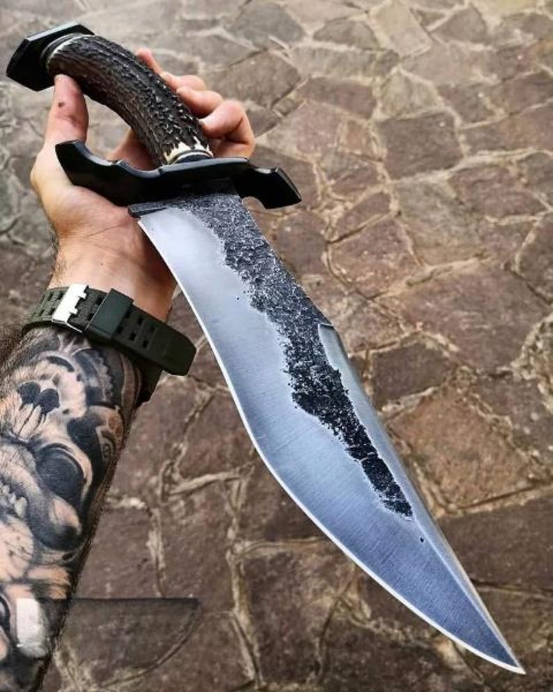 Hand holding a large knife with a textured handle on a stone pavement background
