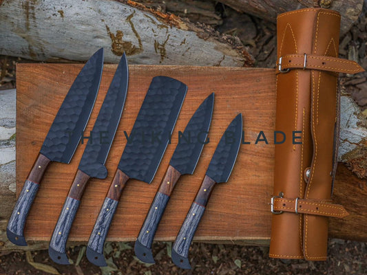 A set of five stainless steel kitchen knives with black coated blades and brown wooden handles, displayed alongside a brown leather roll kit case, placed on a wooden surface with a background of logs.